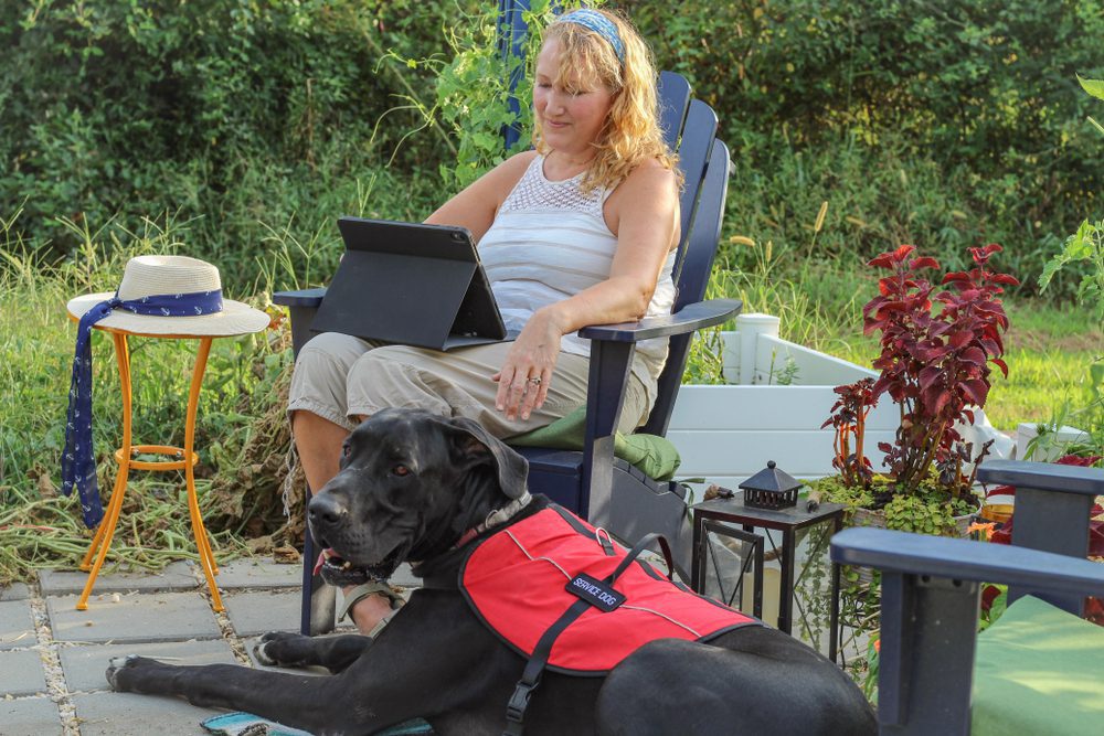 White woman sitting outside with a tablet in her lap and her guide dog sitting beside her.